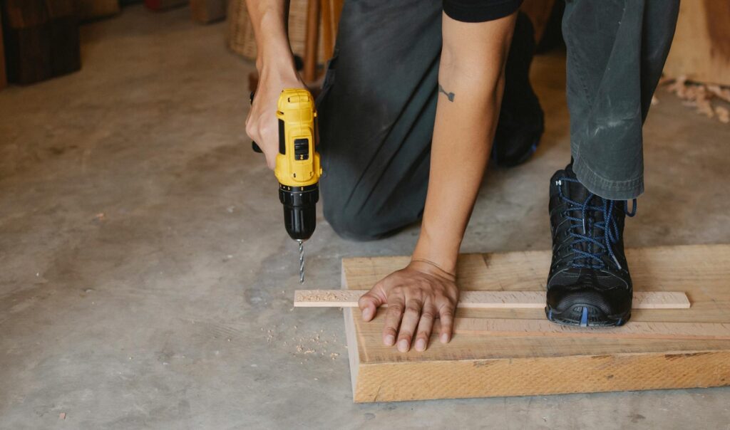 Person using a yellow power drill on wooden boards while wearing work boots.