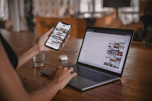 Person viewing a photo gallery on phone and laptop simultaneously on wooden desk.