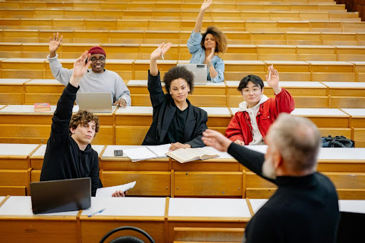 Students raising hands in a lecture hall with tiered wooden seating.